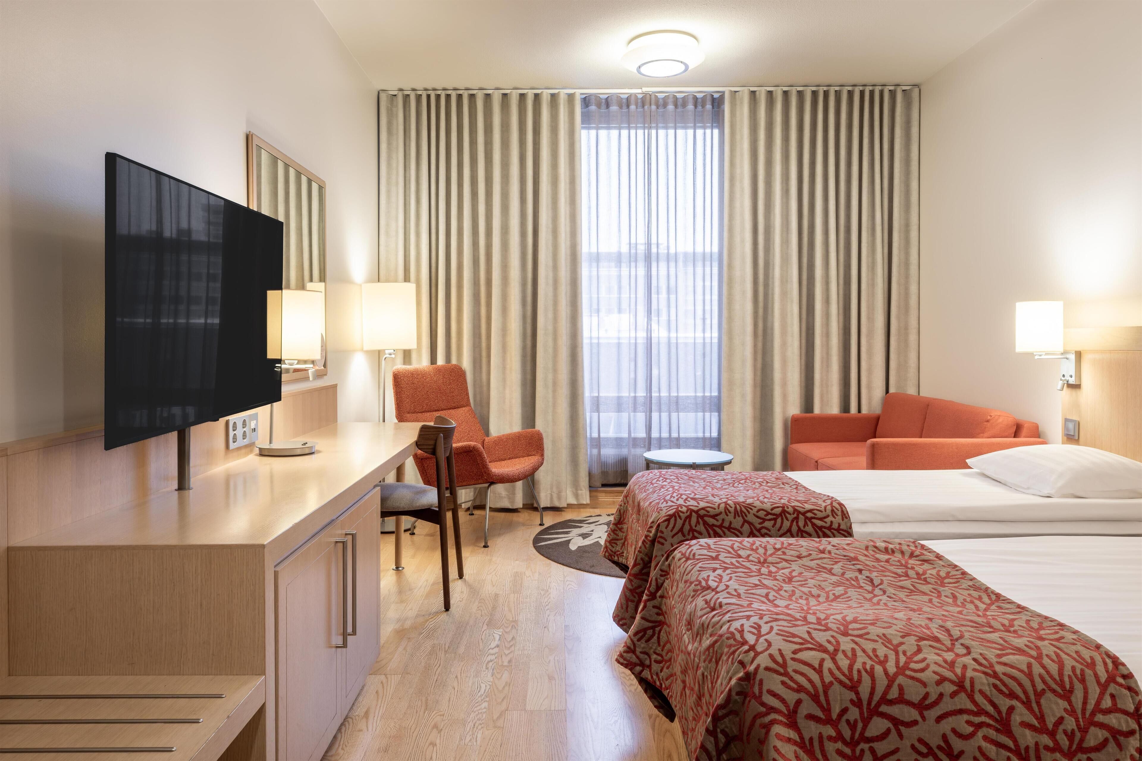 Habitación de hotel con escritorio, televisión, dos camas, sofá y ventana con cortinas.