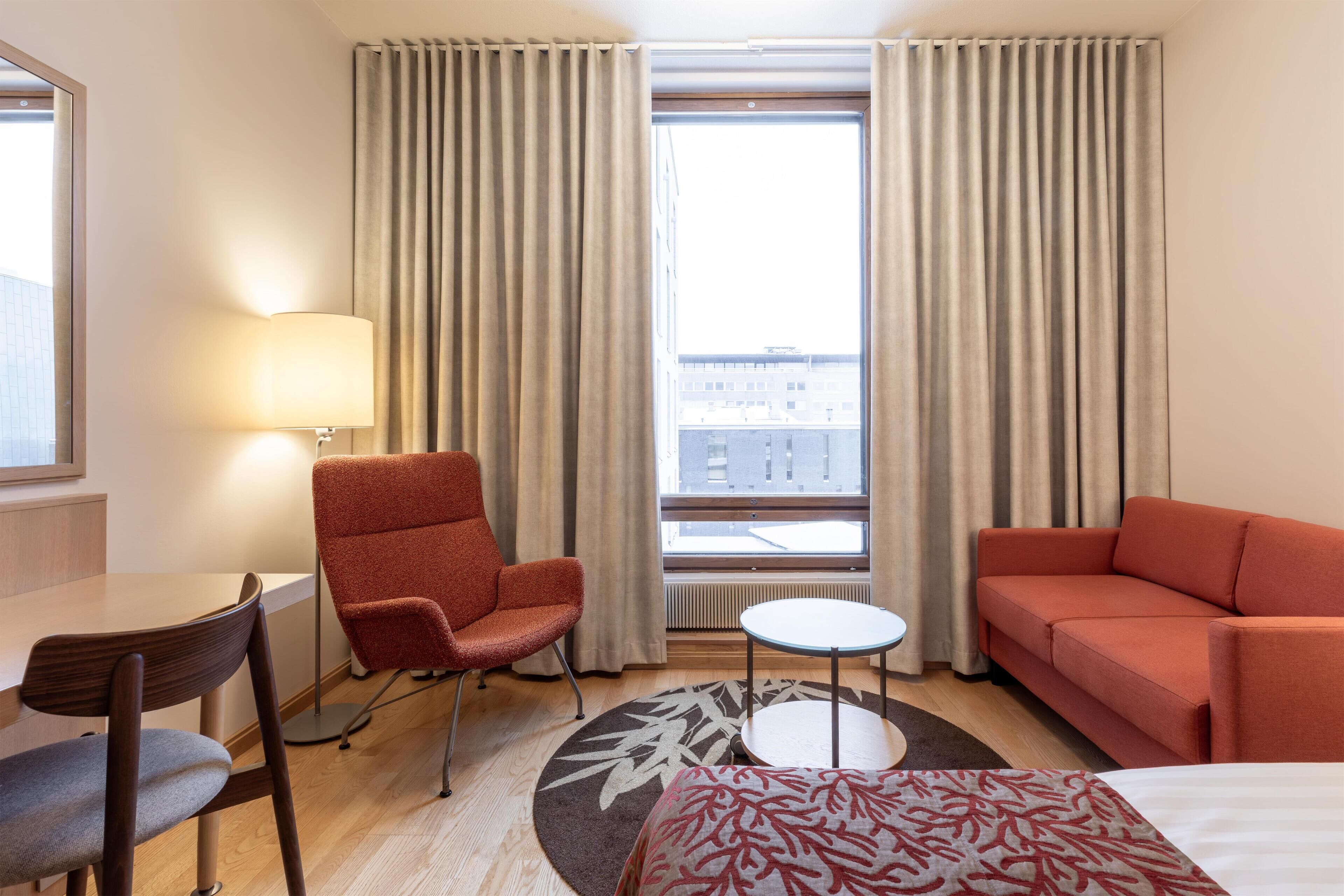 A hotel room with a bed, a red armchair, a small round table, a sofa, and a lamp.