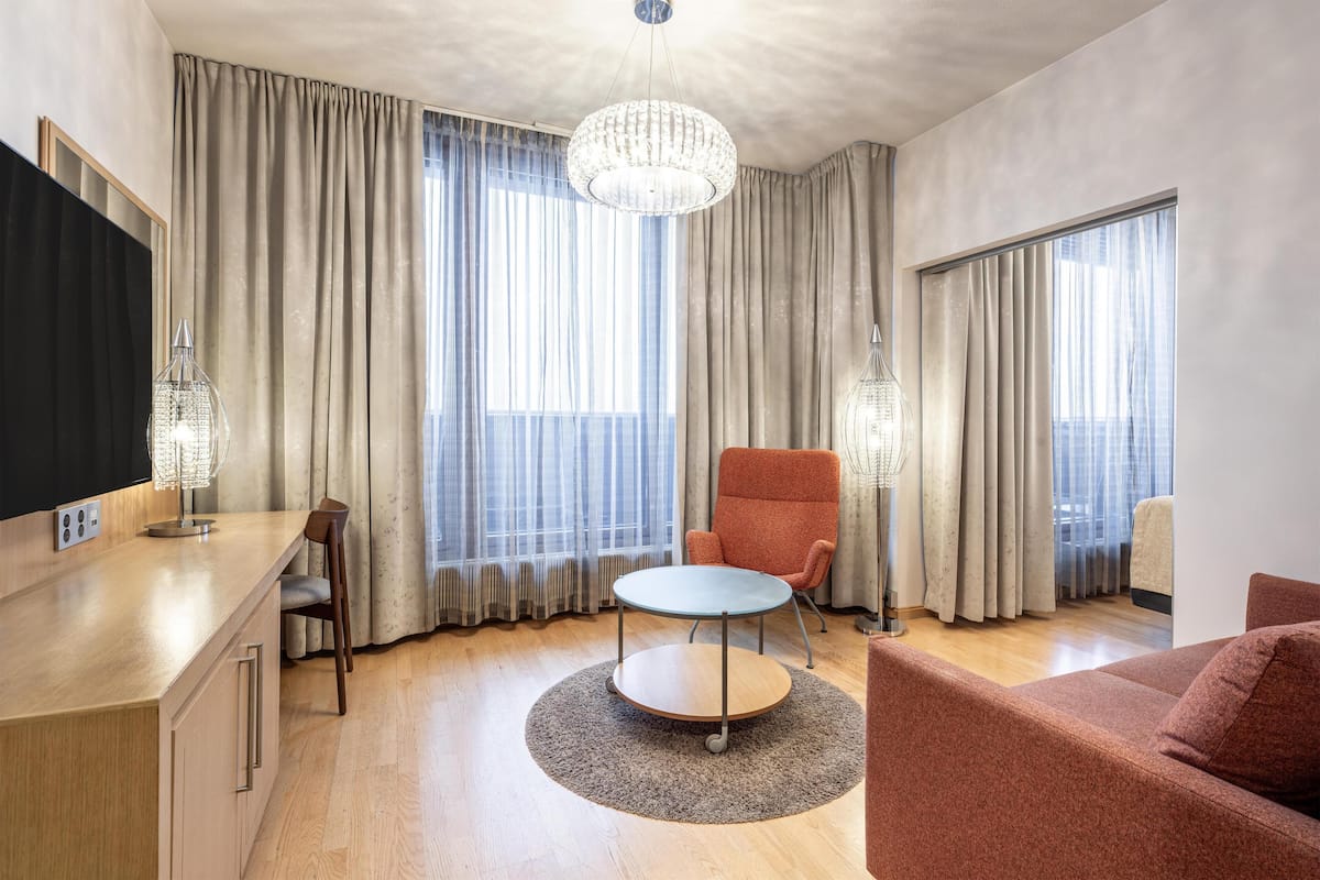 Ein modernes Wohnzimmer mit Holzboden, einem runden Tisch, einem roten Sessel, einem beigen Sofa und einem großen Fenster mit Vorhängen.