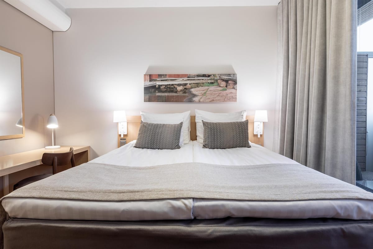 Ein Hotelzimmer mit einem großen Bett, zwei Nachttischen inklusive Lampen, einer Kopfteil mit Stadtbild-Mural und Blick ins Freie durch ein Fenster mit Vorhängen.