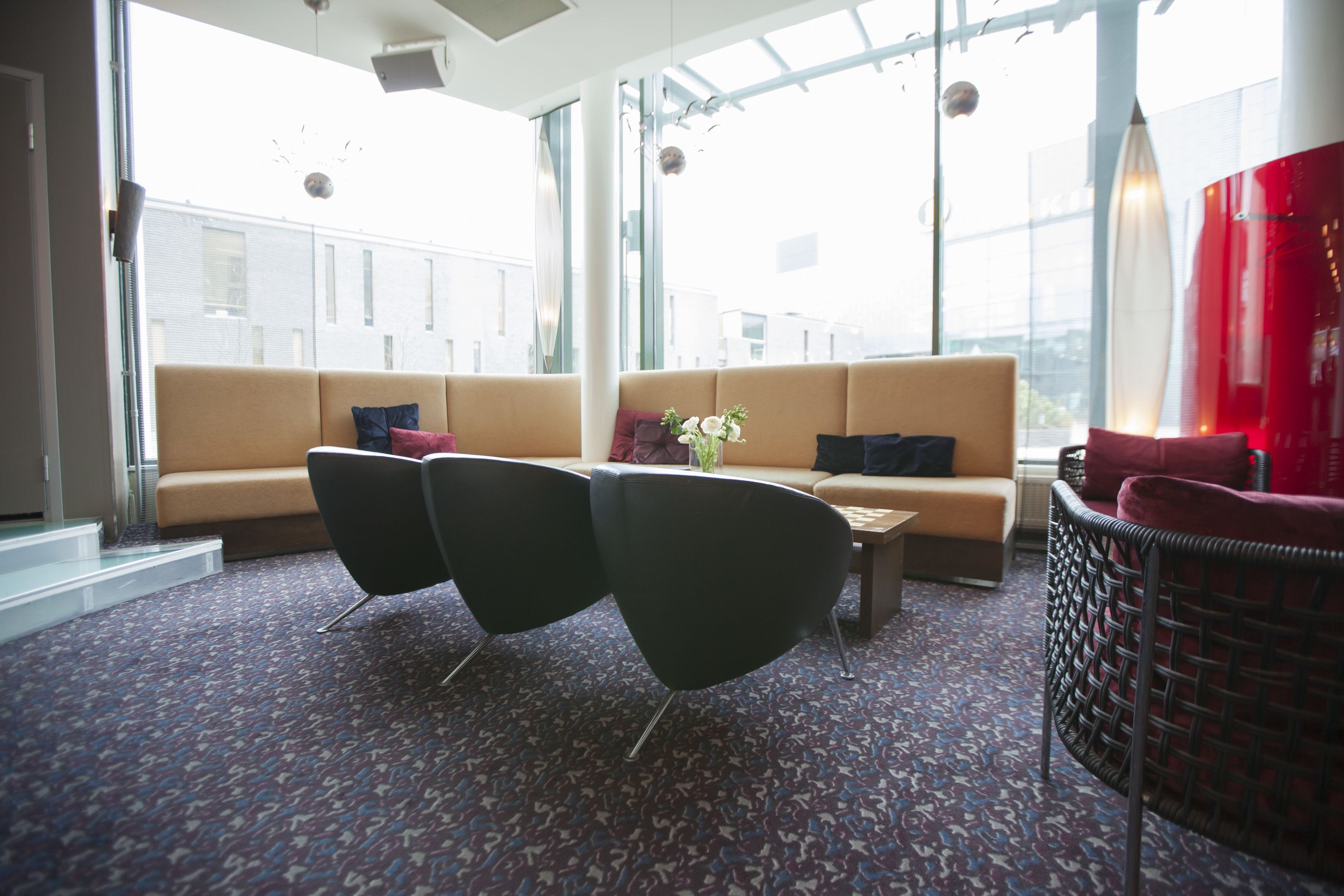 Un'area lounge moderna con un divano, due poltrone nere, un tavolino e una poltrona in vimini. Ampie finestre offrono vista sulla città.