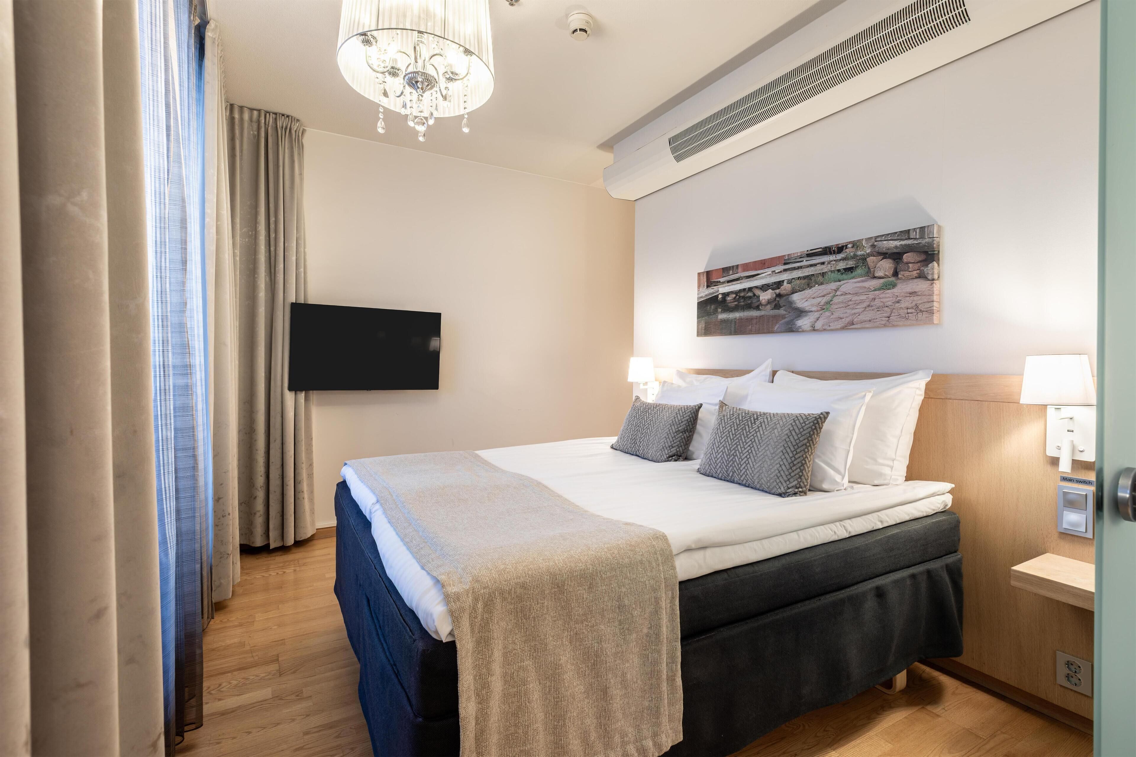 A hotel room with a bed, a flat-screen TV mounted on the wall, a chandelier, and a painting above the bed.