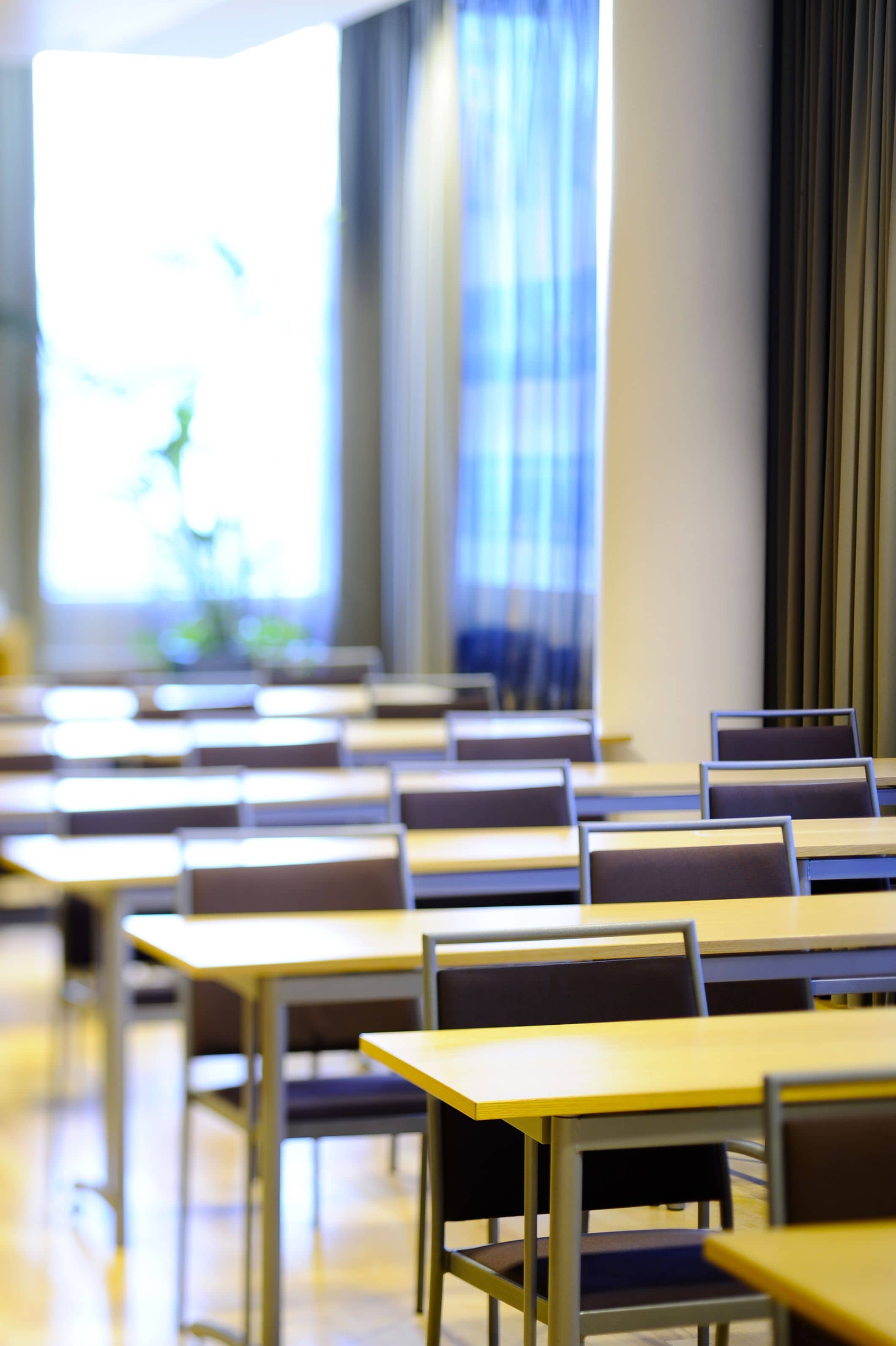 Ein Klassenzimmer mit Tischen und Stühlen, großen Fenstern mit Vorhängen und hellem Innenraum.