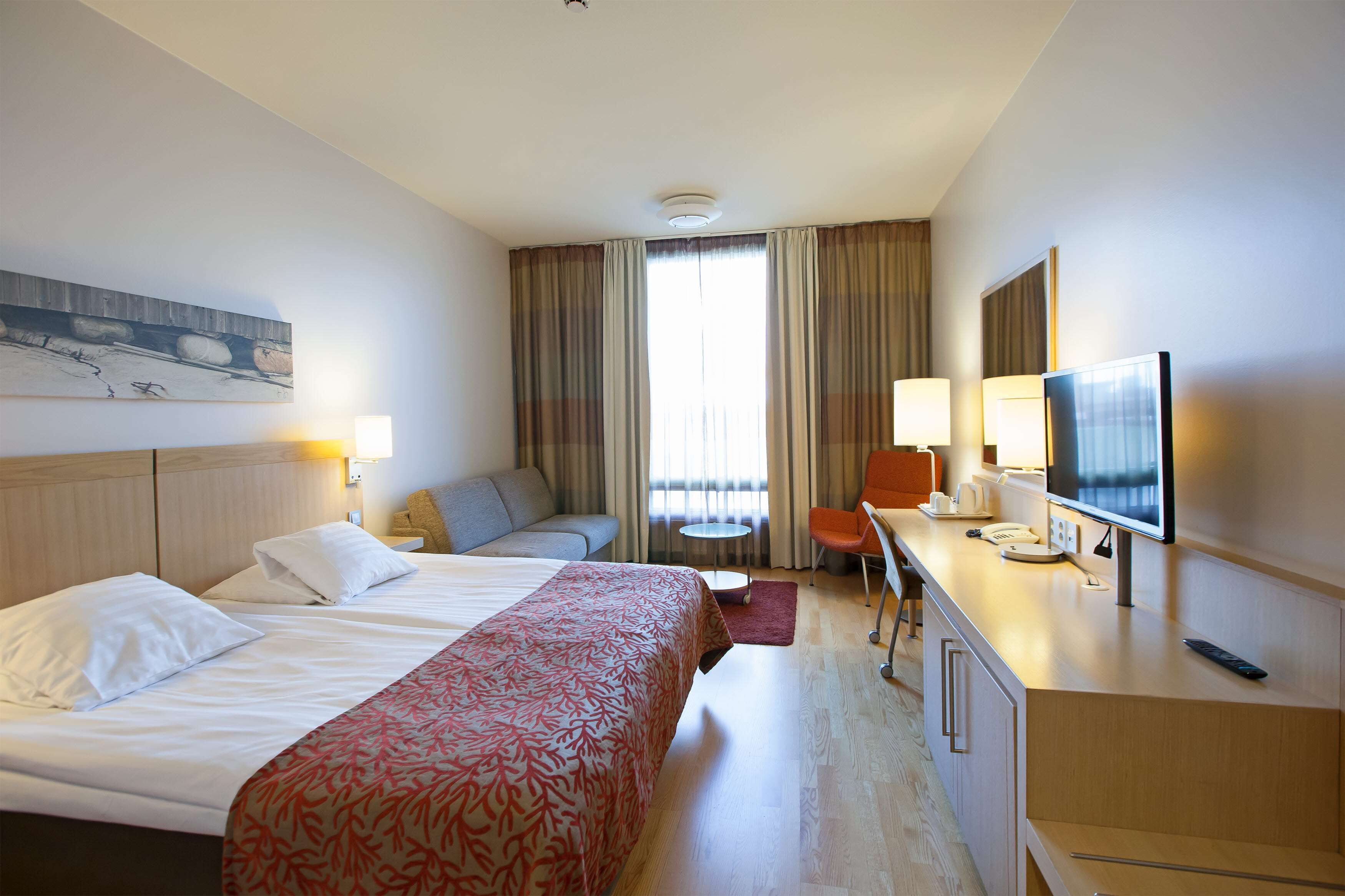 Habitación de hotel con dos camas, un sofá, un escritorio con televisor y una ventana con cortinas.