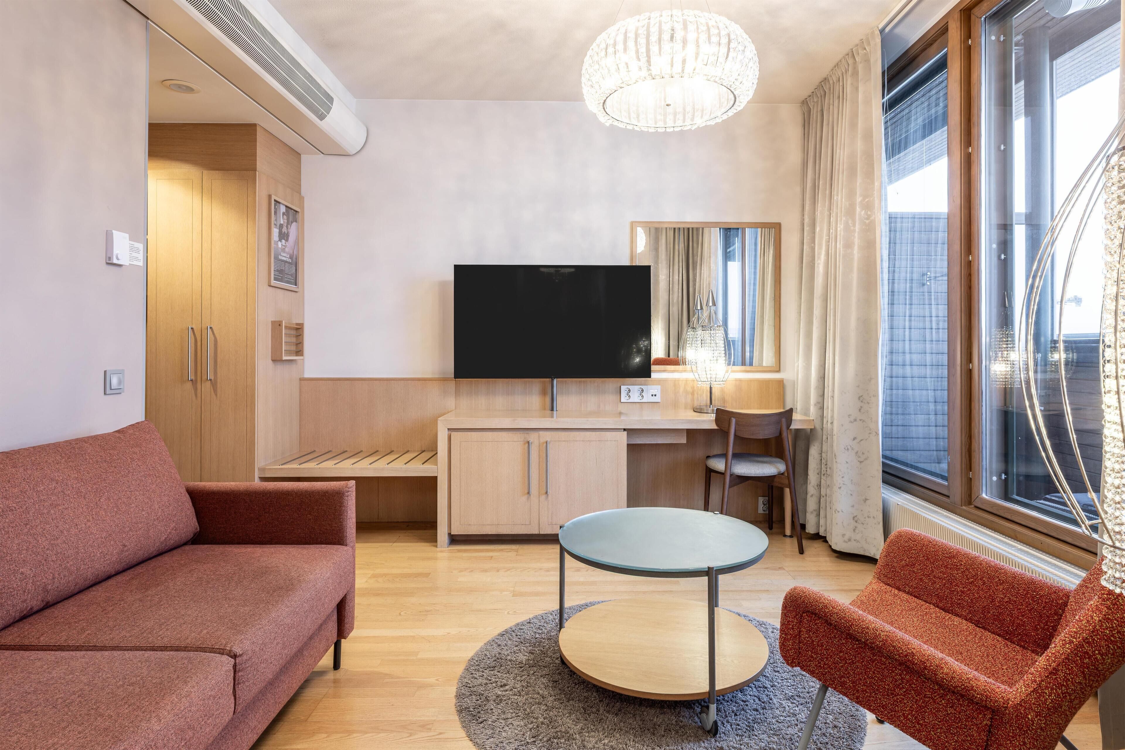A modern hotel room with a sofa, a round table, a TV, a desk, and a large window.