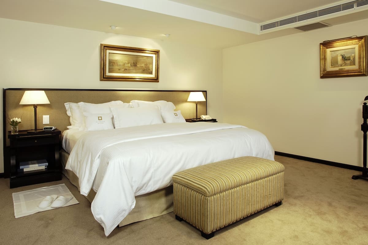 Une chambre d’hôtel avec un grand lit, deux tables de chevet avec des lampes, une petite table de chevet avec un téléphone, un cadre photo fixé au mur, une lampe sur pied et un banc.