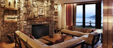 A cozy living room with a stone fireplace, leather sofas, and a large window overlooking a body of water.