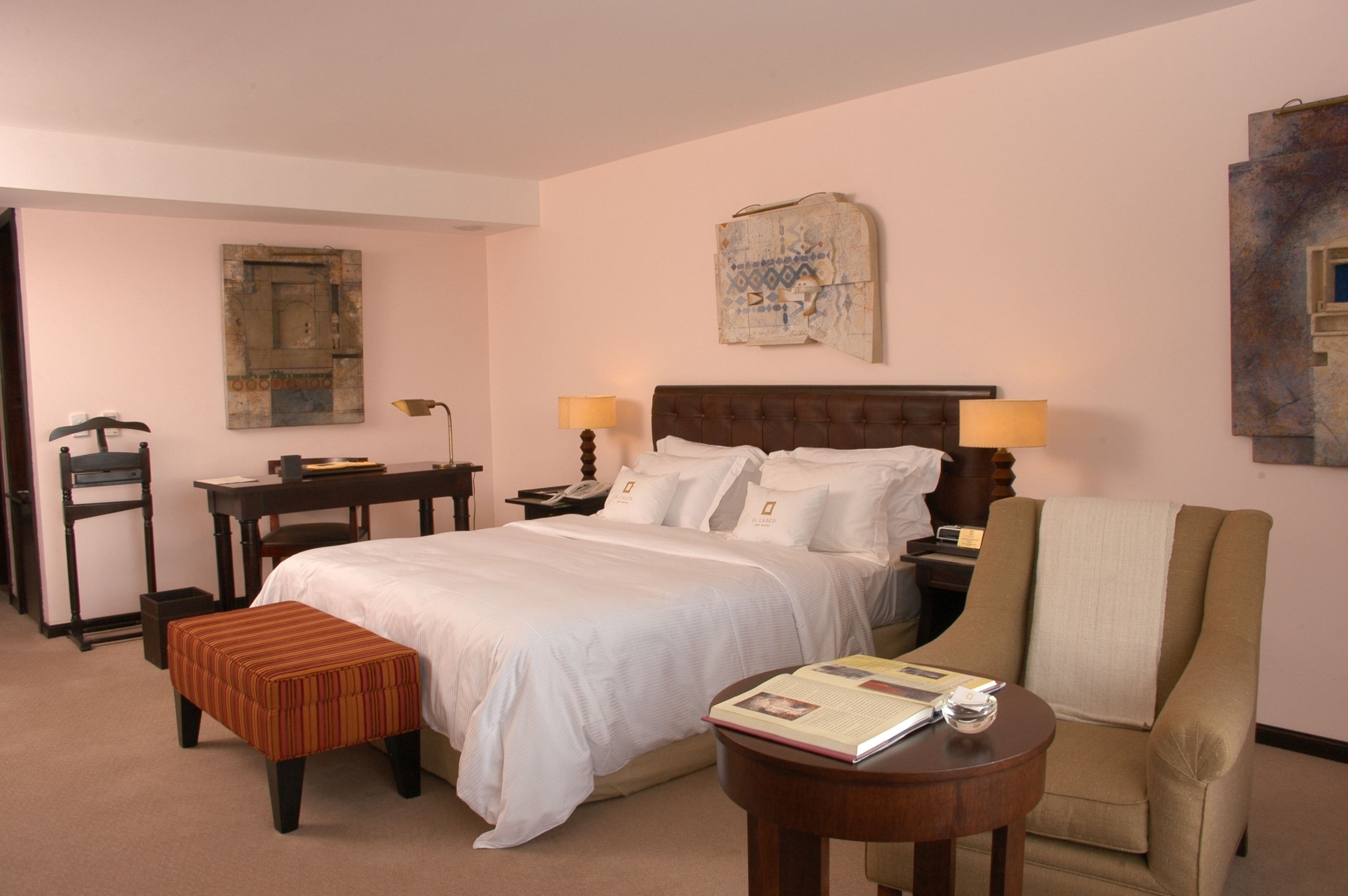 A hotel room with a large bed, a desk, a chair, and a small table with a book.
