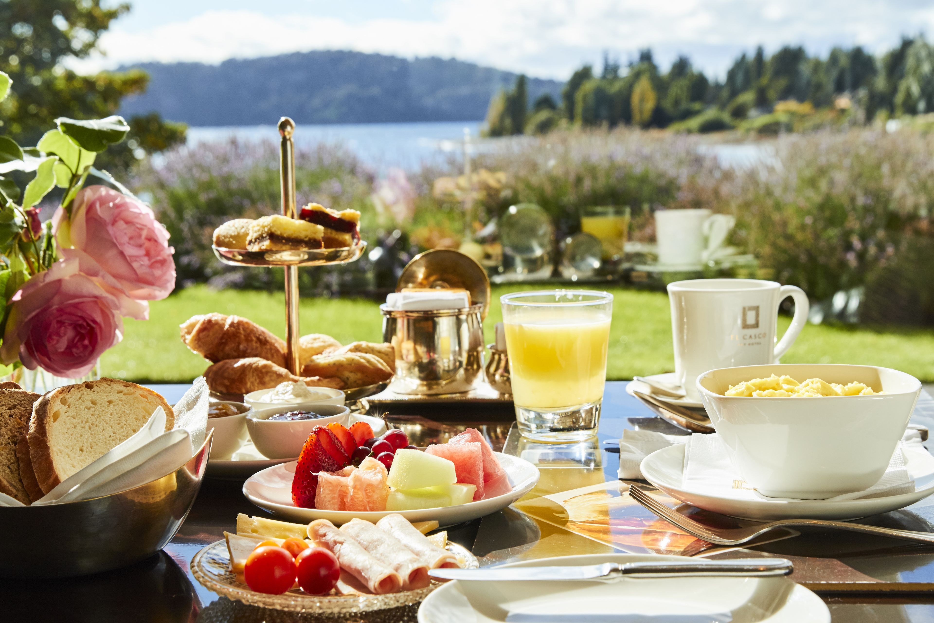 A breakfast table with various food items, a cup of coffee, and a scenic view of a lake and hills in the background.