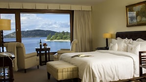 A hotel room with a large window offering a view of a lake and forest. There is a bed with white linens, a bedside table with a lamp, and a chair. - El Casco Art Hotel (San Carlos de Bariloche)