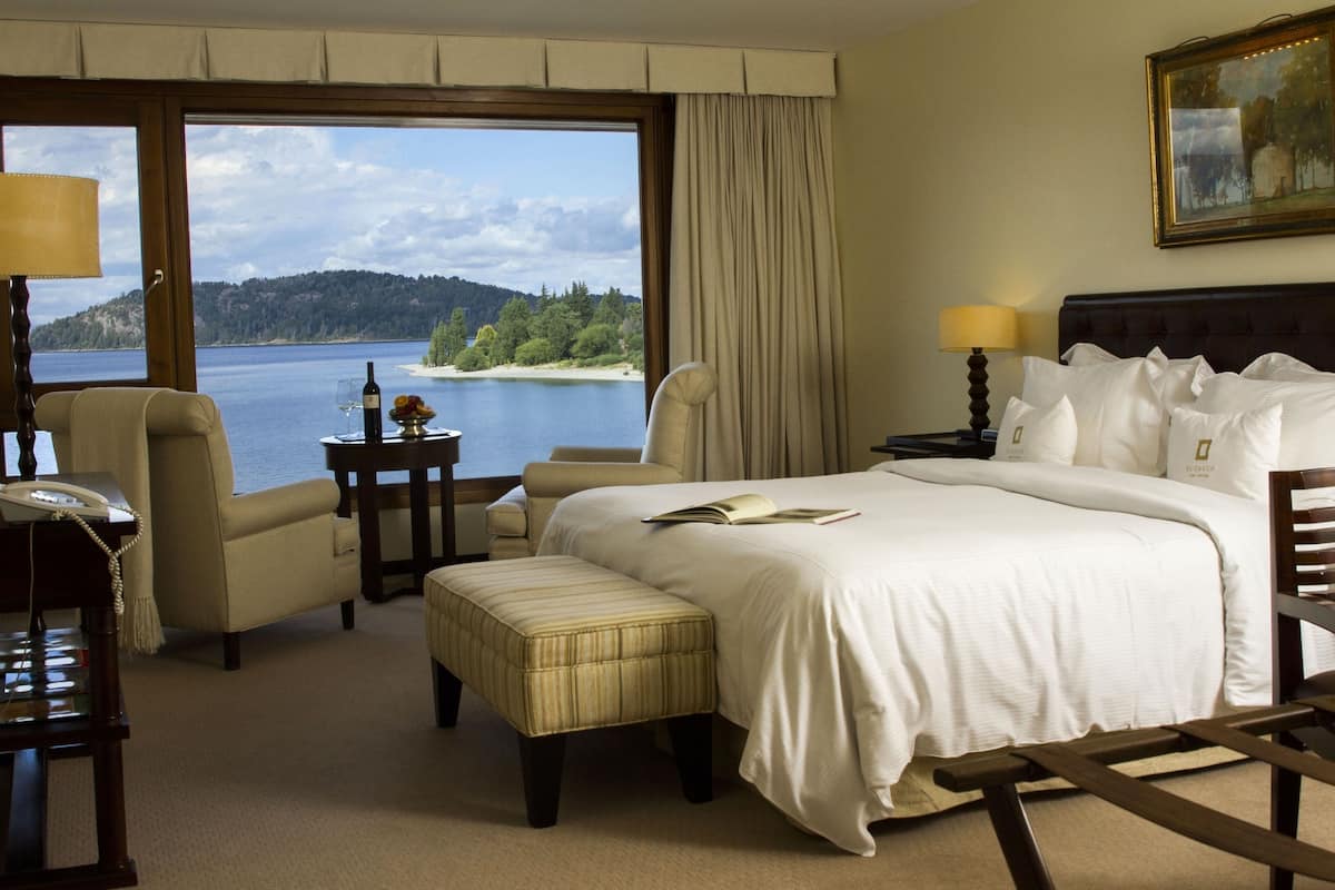Une chambre d’hôtel dotée d’une grande fenêtre donnant sur un lac et une forêt. On y trouve un lit avec du linge de maison blanc, une table de chevet avec une lampe et un fauteuil.
