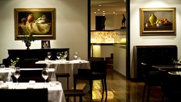 Un espace repas avec des tables préparées pour deux personnes, des œuvres d’art encadrées accrochées aux murs et une vue sur une autre pièce.