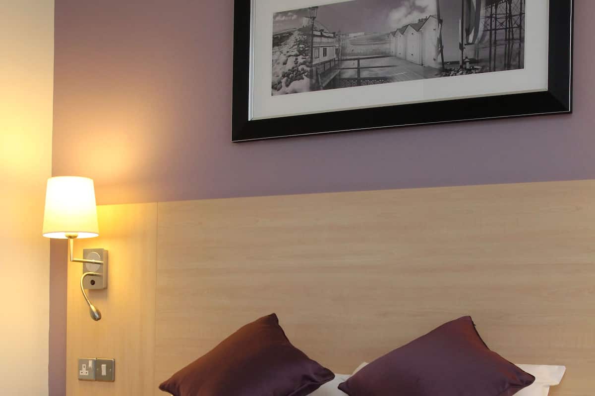 A hotel room with a neatly made bed, a framed picture on the wall, and a bedside lamp.