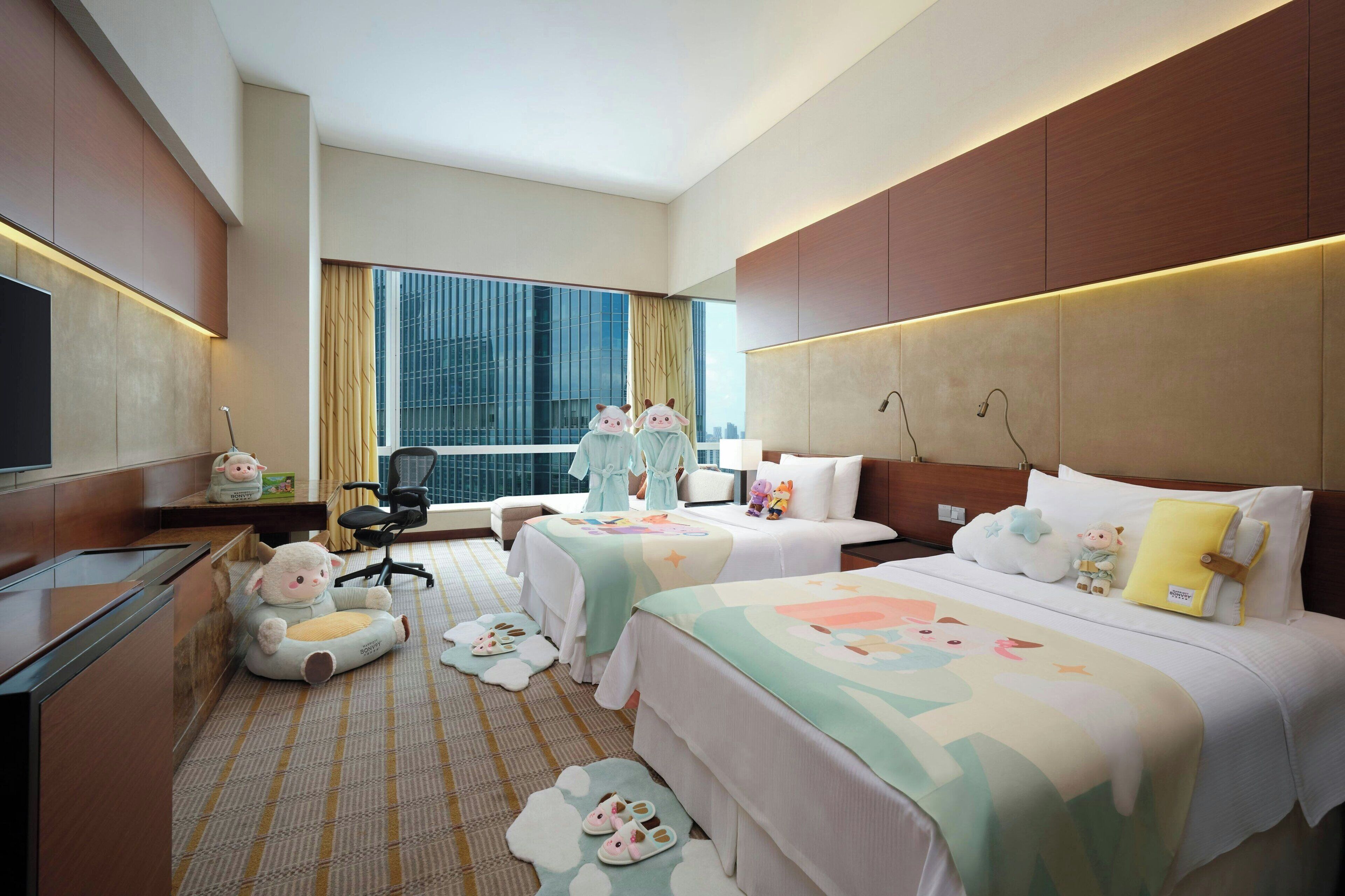 A hotel room with two beds, a desk, a TV, and a large window with a city view.