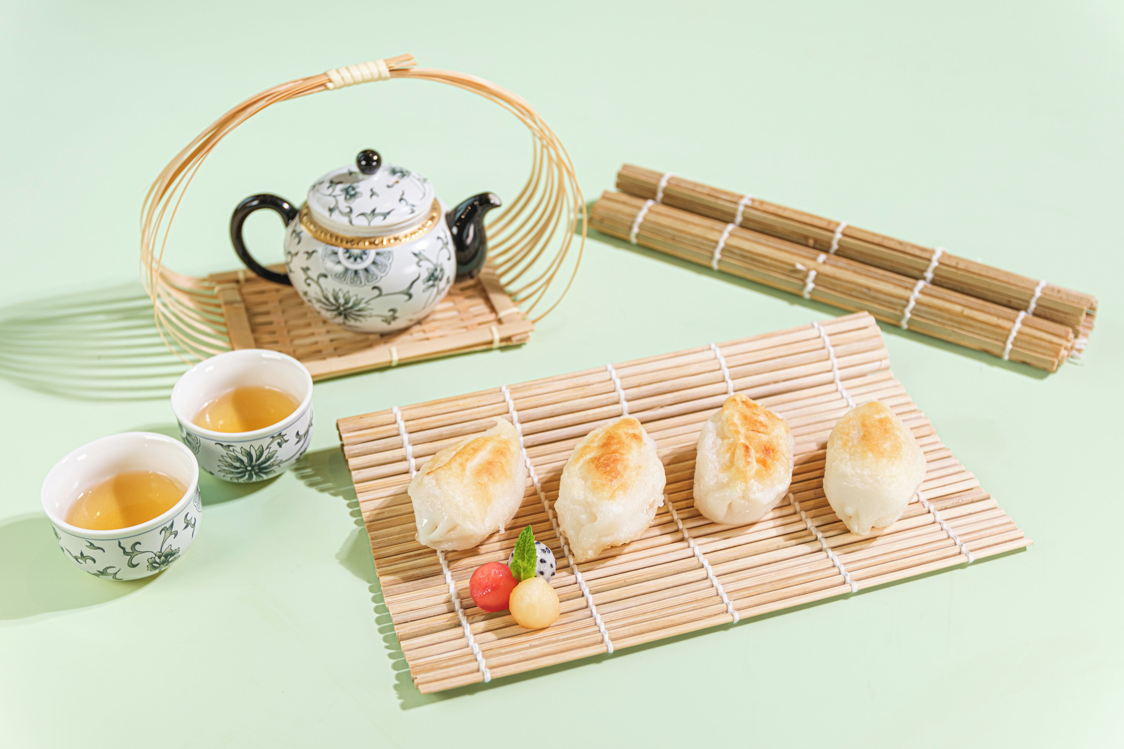A tea set with a teapot and cups, a bamboo mat with dumplings, and a small fruit arrangement.