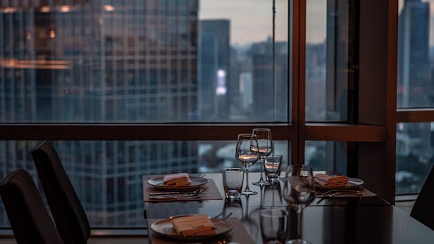 A dining table set for two with a cityscape view through large windows.