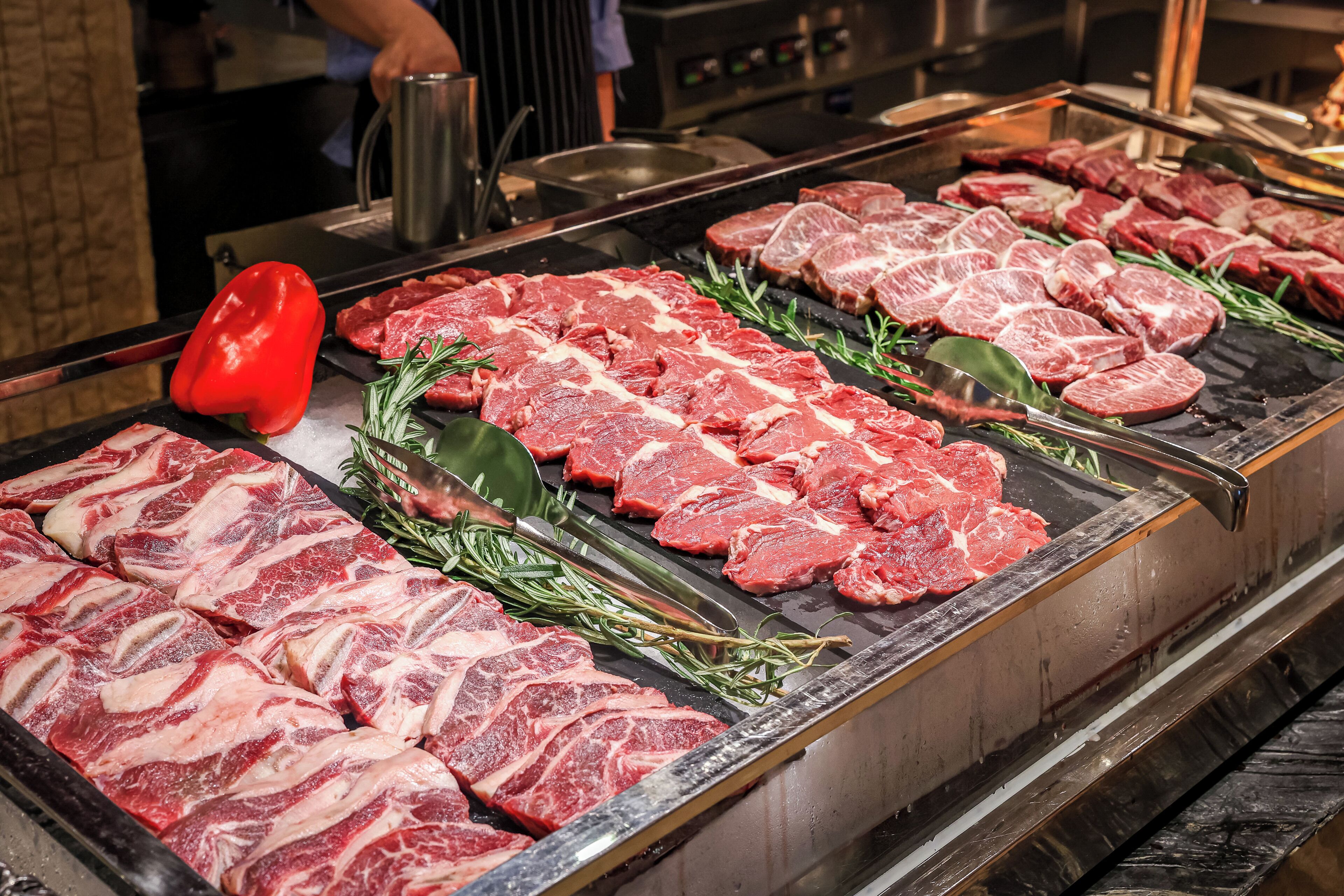 Exhibición de diferentes cortes de carne cruda, incluyendo ternera y cerdo, dispuesta sobre una superficie metálica, con un pimiento rojo y ramitas de romero como decoración.