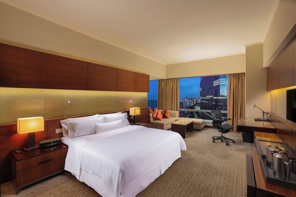 A hotel room with a large bed, a desk, a chair, and a view of a cityscape through the window.