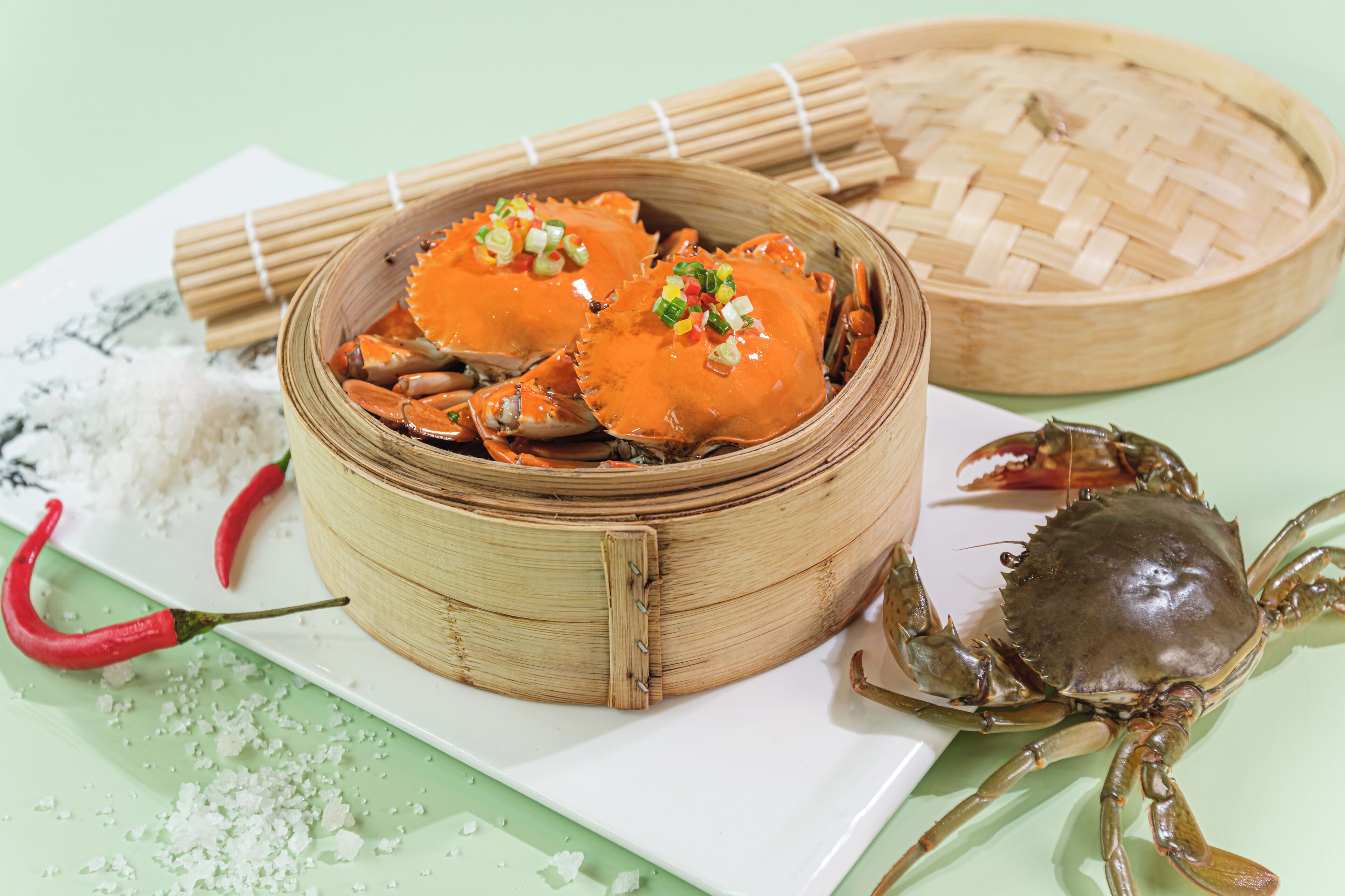 A bamboo steamer with cooked crabs, a whole crab, and a red chili pepper on a white plate.