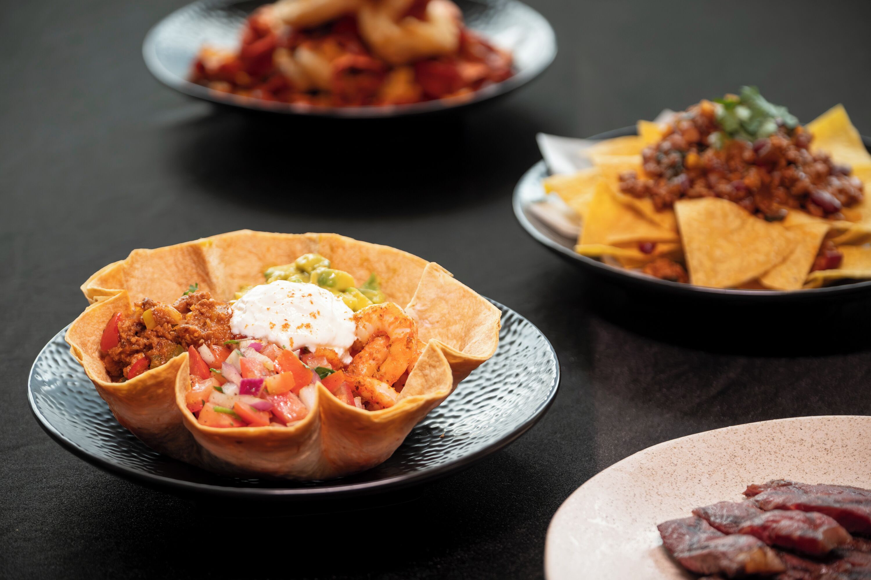 Three plates of food, including tacos and nachos, on a dark table.