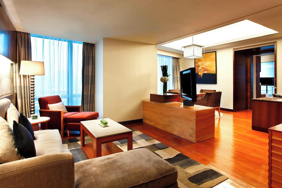 A modern hotel room with a sofa, armchair, and a small table. There is a TV and a wooden desk in the corner.