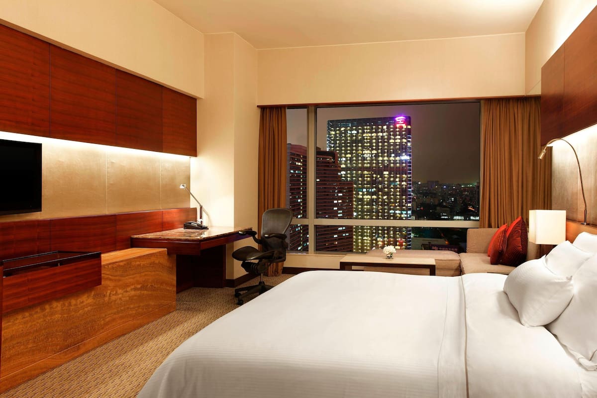 A hotel room with a large bed, a desk, a chair, a TV, and a view of a cityscape at night.