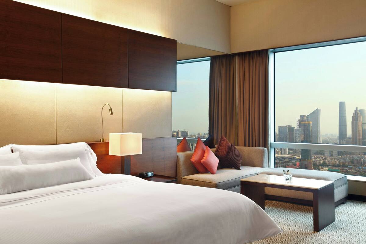 A hotel room with a large bed, a sofa, a coffee table, and a view of a cityscape through a large window.