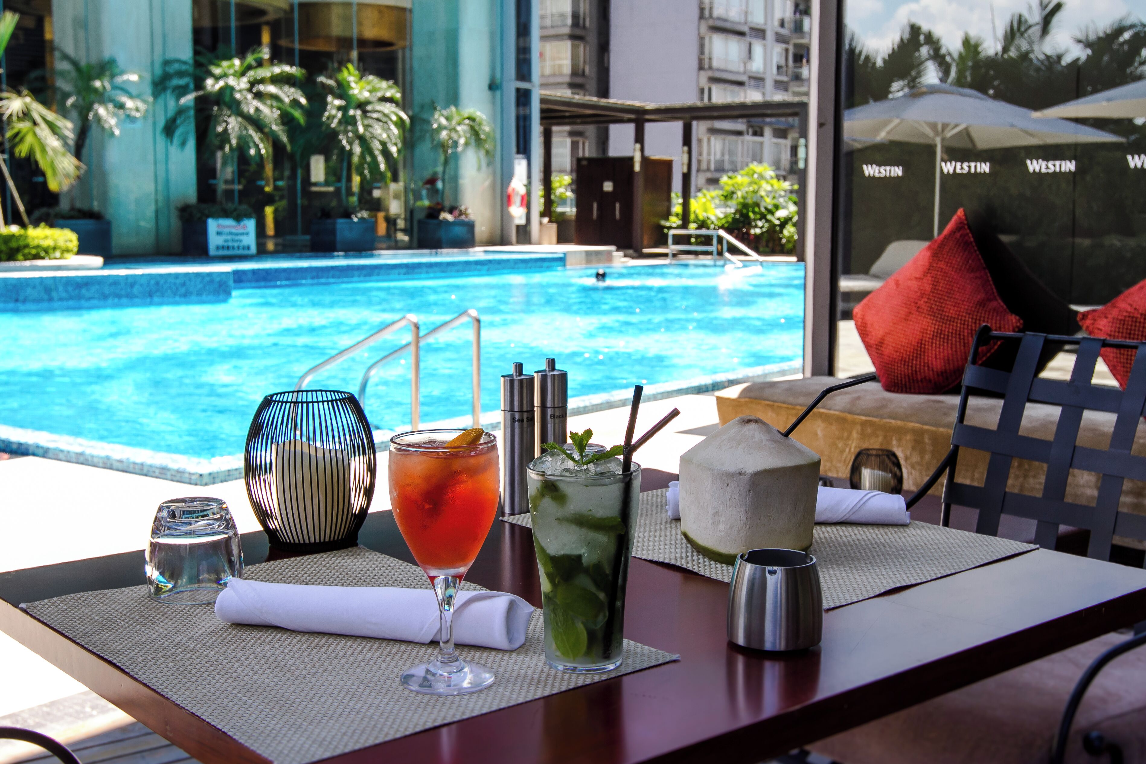 A poolside table with drinks and a coconut, overlooking a swimming pool.