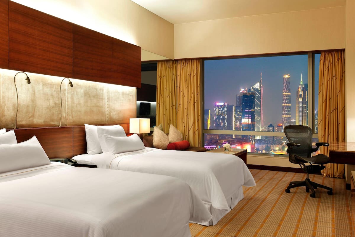 A hotel room with two beds, a desk, and a chair. The room has a large window with a cityscape view.