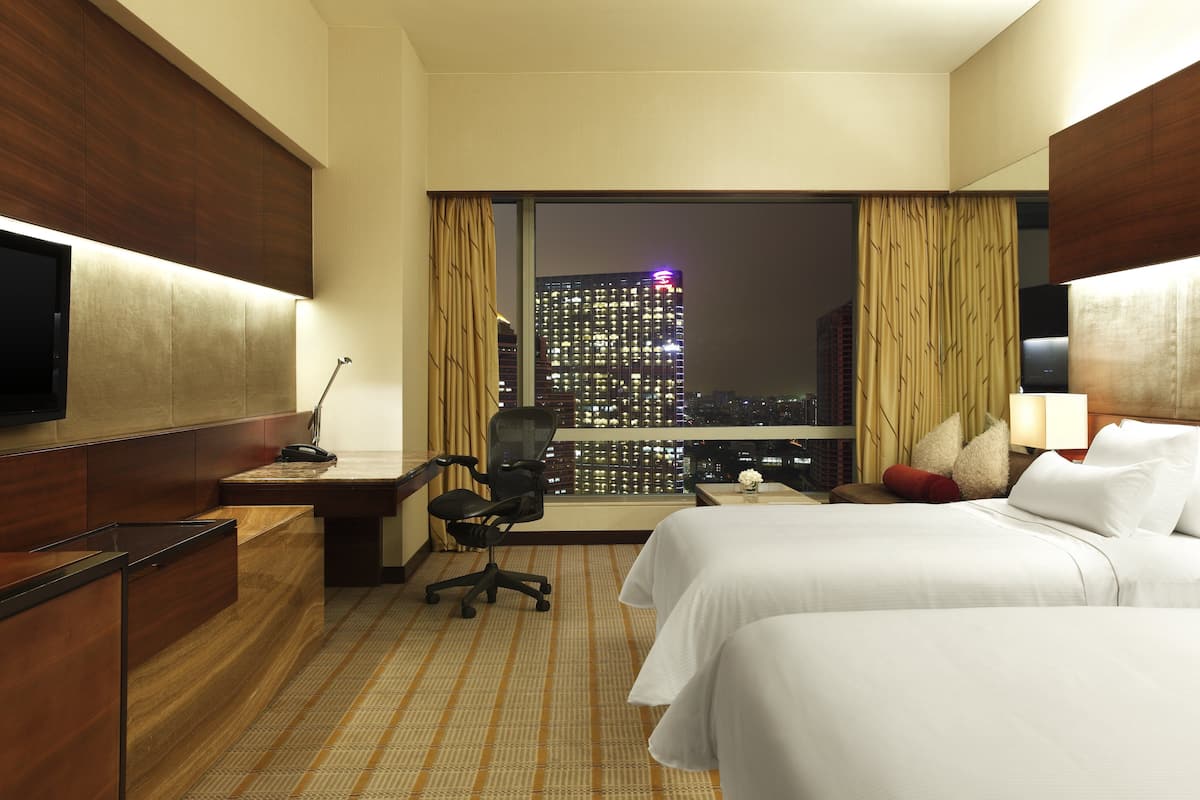 A hotel room with two beds, a desk, a chair, a TV, and a large window with a city view.
