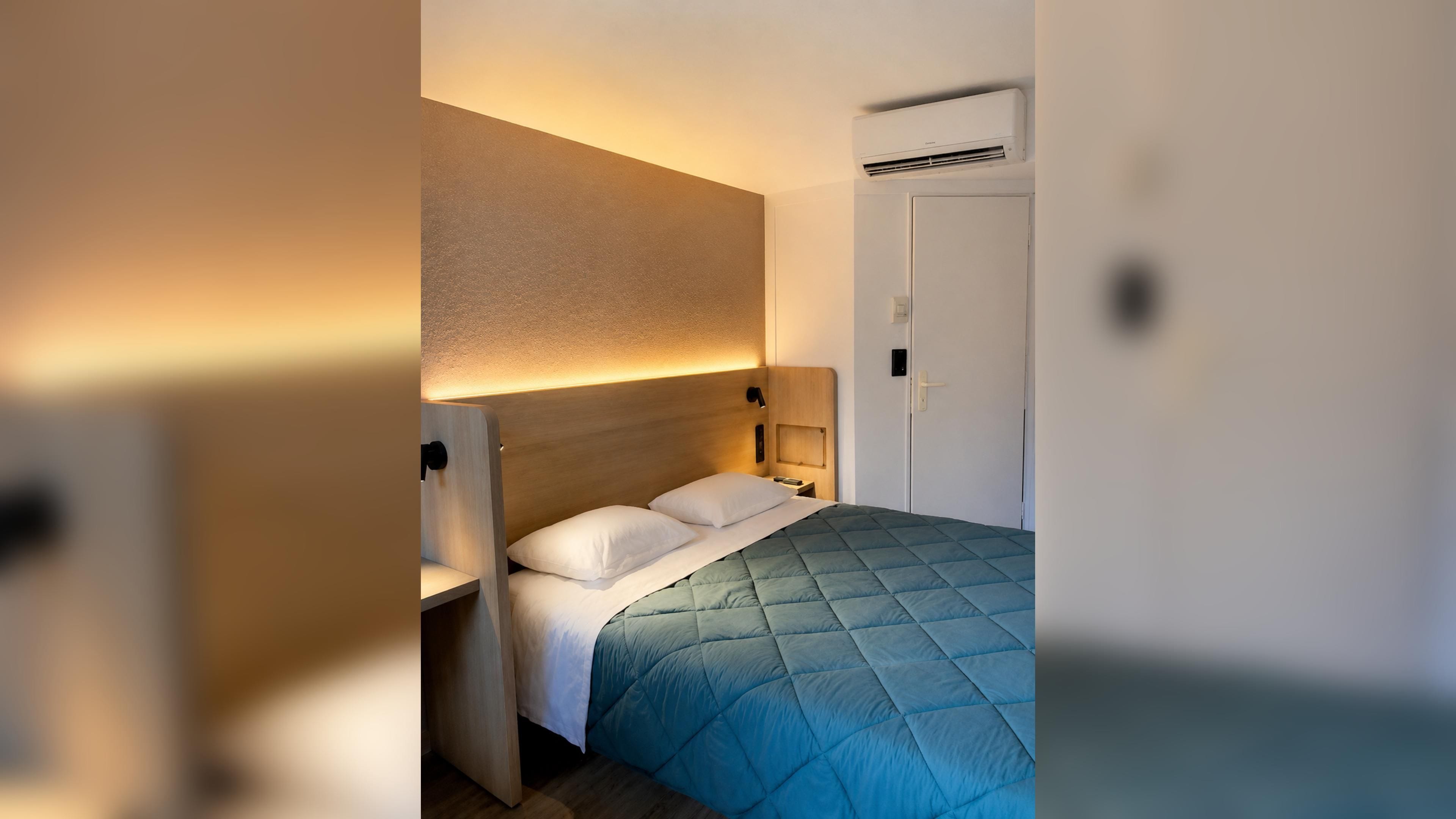 A hotel room with a bed, bedside table, wall-mounted air conditioning unit, and a door.