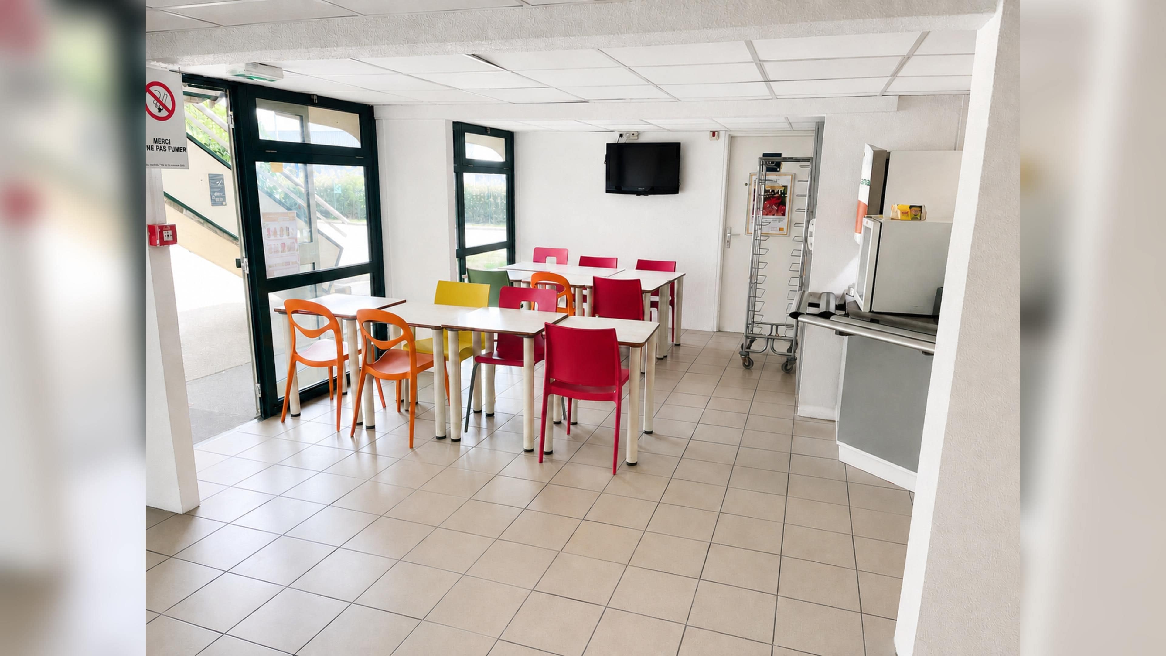 A clean, brightly lit indoor space with tables and chairs arranged for dining, a television mounted on the wall, and a door with a sign.