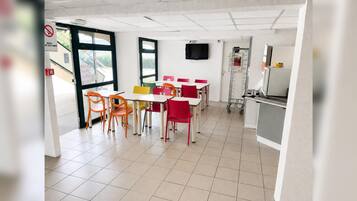 A clean, brightly lit indoor space with tables and chairs arranged for dining, a television mounted on the wall, and a door with a sign.
