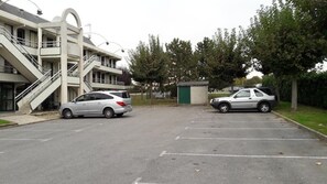 Un estacionamiento con dos coches, un edificio con un diseño arquitectónico singular que incluye una escalera en espiral, y una pequeña estructura verde al fondo.