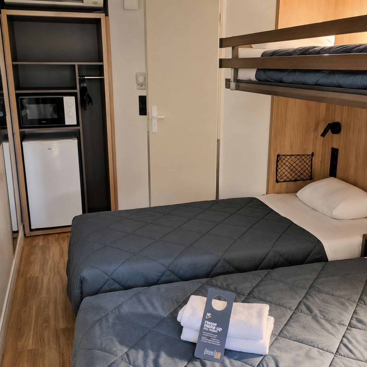 A hotel room with a bunk bed, a small kitchenette, and a bedside table with a towel.