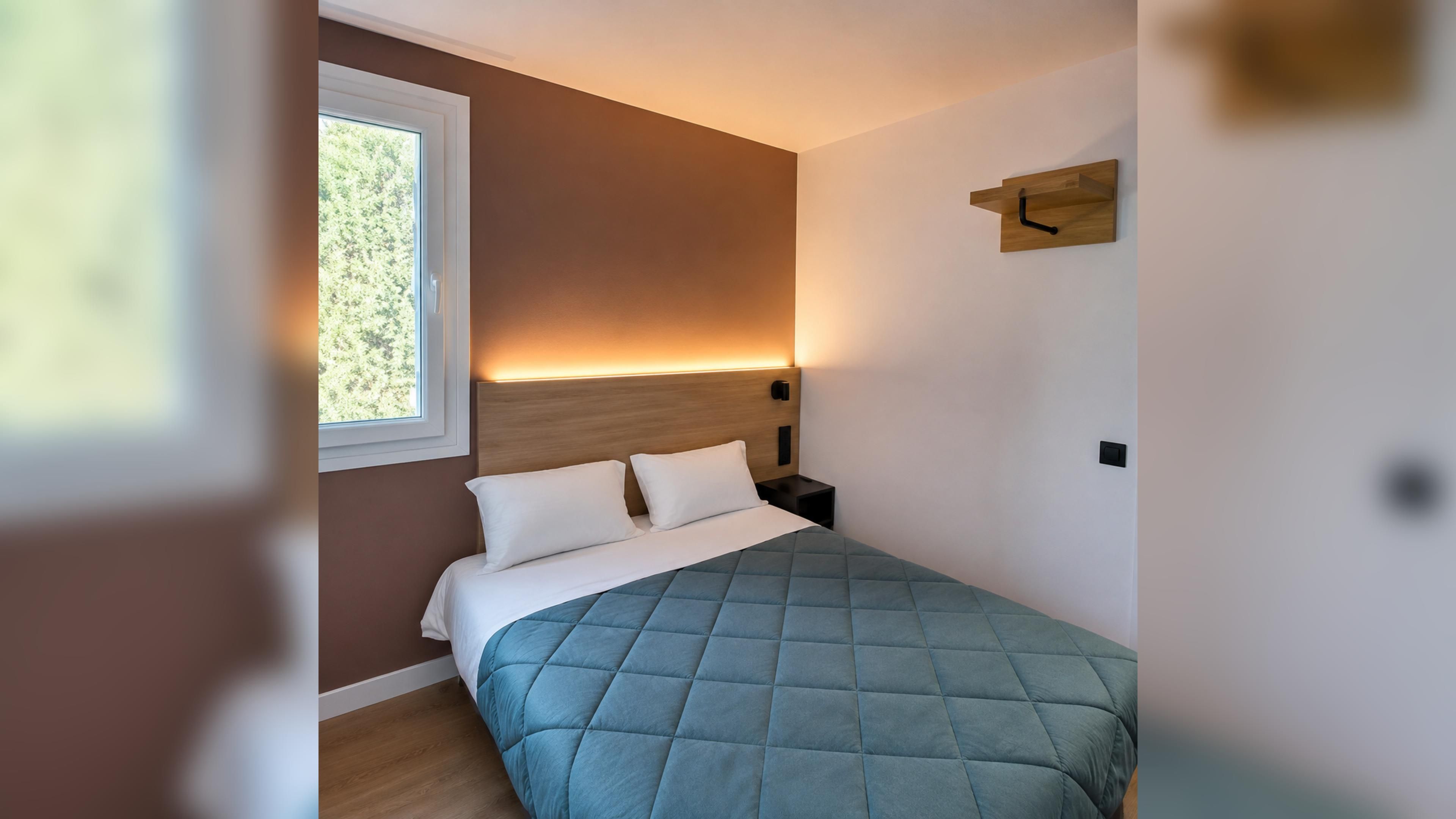 A bedroom with a bed, a nightstand, a wall-mounted clock, and a window with a view of trees.
