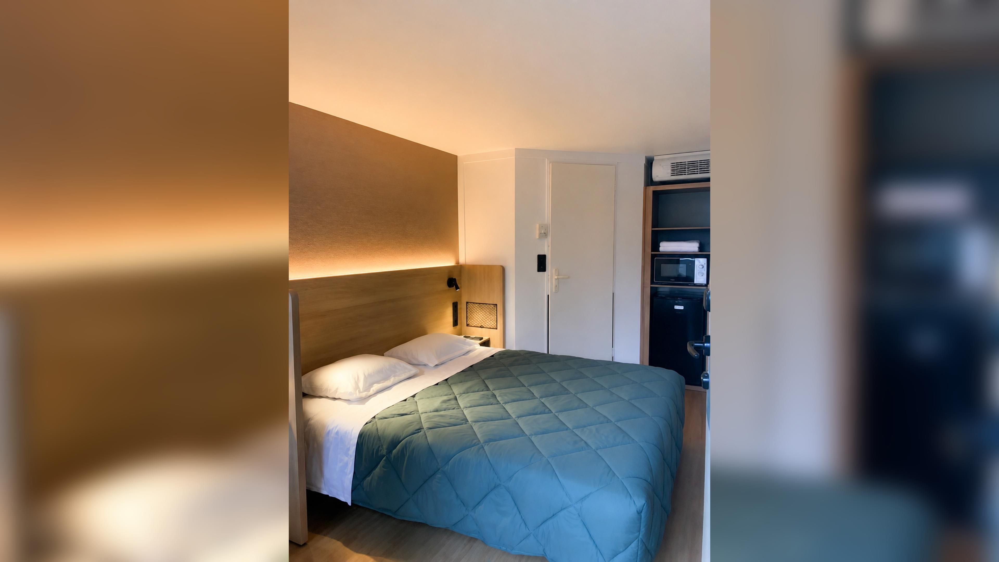 Een hotelkamer met een bed, nachtkastje en een kledingkast met een magnetron en een klein apparaat.