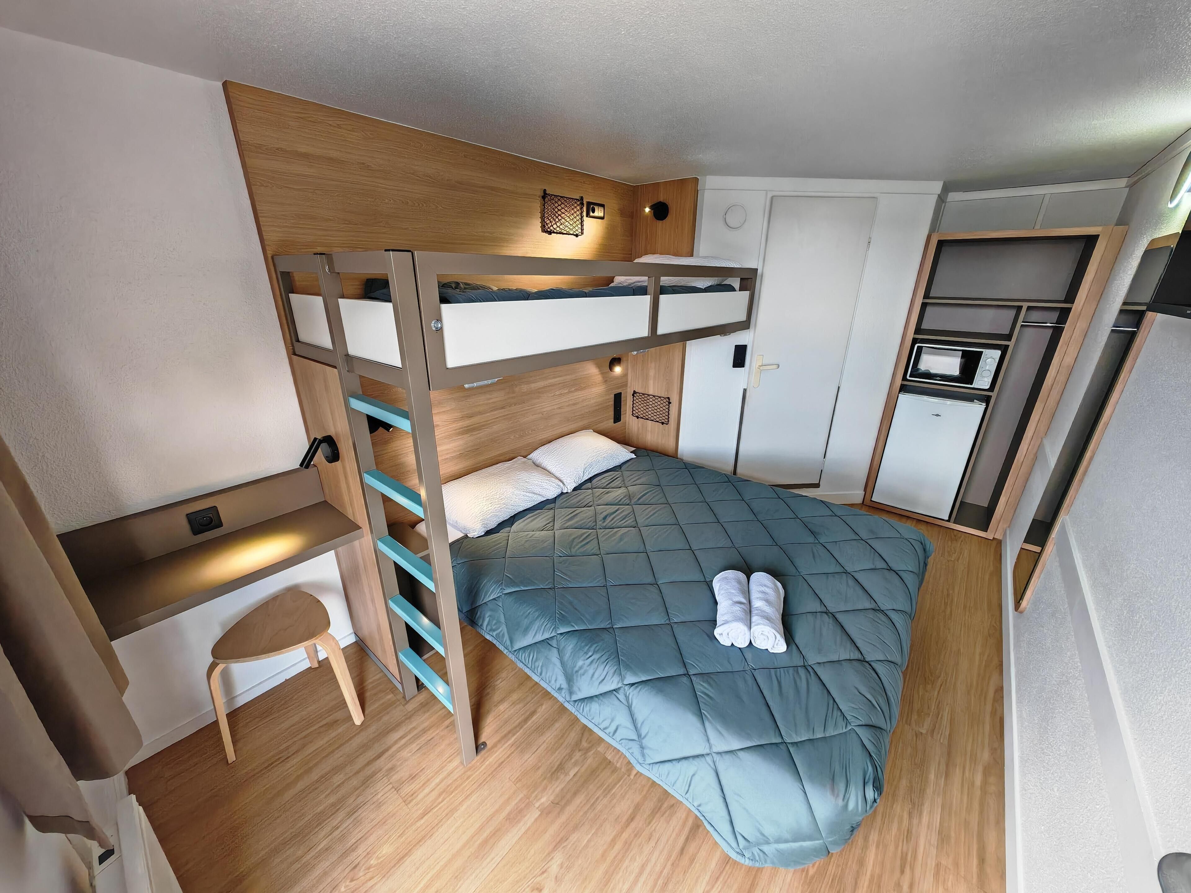Una cama en literas con escritorio y silla, una kitchenette pequeña y un armario.