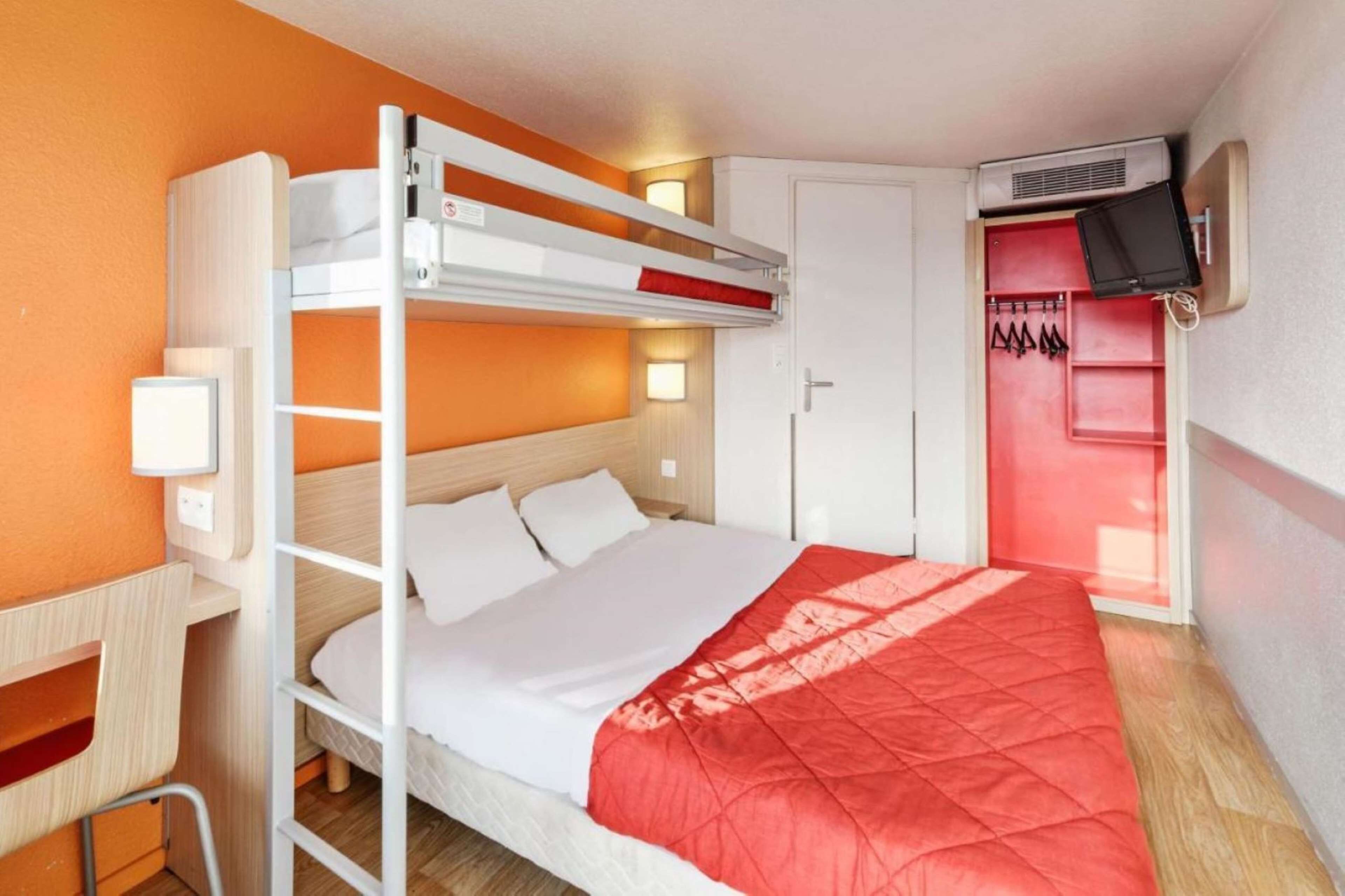 A bunk bed room with a red blanket, a desk, a chair, a wardrobe, and a TV mounted on the wall.