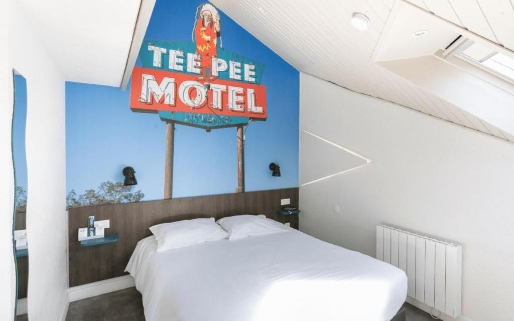 Een hotelkamer met een bed, een nachtkastje, een wandlamp en een bord waarop staat 'TEE PEE MOTEL'.