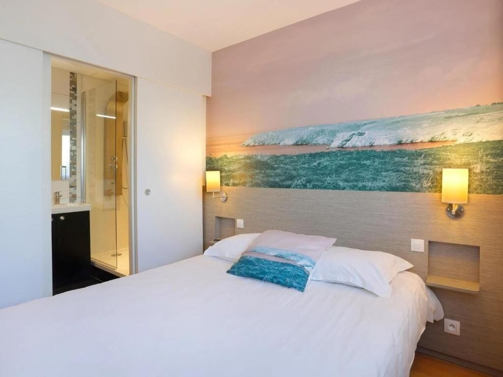 A hotel room with a bed, a painting of a beach scene, and two wall-mounted lamps.