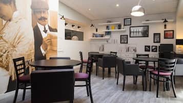 Een moderne eethoek met ronde tafels en zwarte stoelen, een grote muurschildering en planken met boeken en decoratieve objecten.