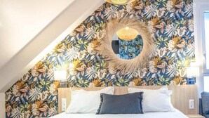 Un dormitorio con techo inclinado, cama con ropa de cama blanca, adorno circular en la pared y papel tapiz estampado con motivos florales.