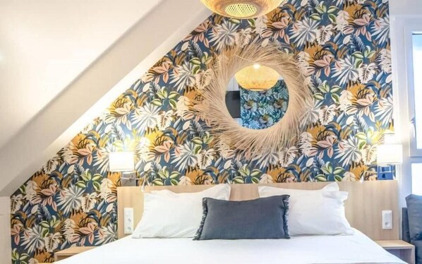 A bedroom with a sloped ceiling, a bed with white linens, a circular wall decoration, and a floral-patterned wallpaper. - Logis Hôtel Au Chêne Vert (Plerin)