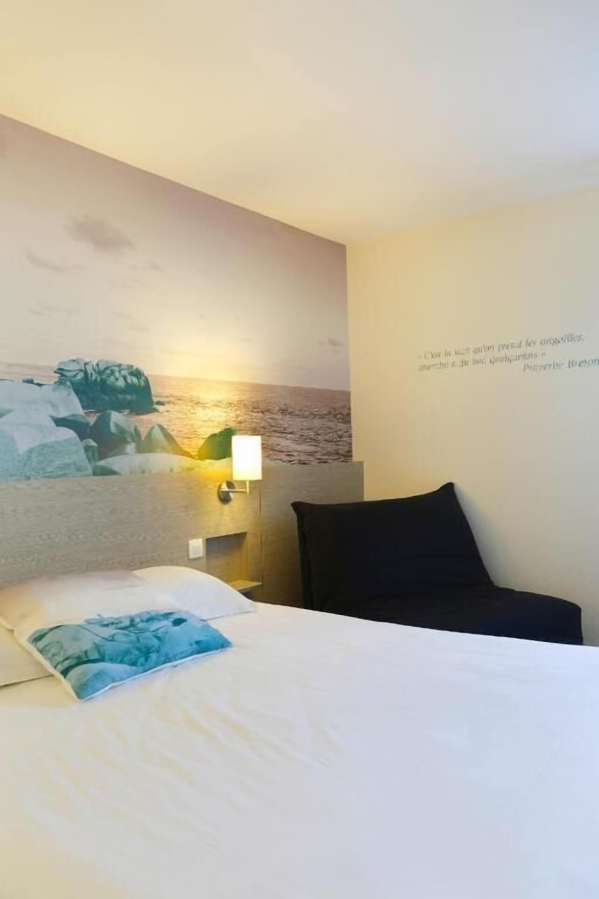 Una camera d'albergo con un letto, una lampada a muro e un quadro raffigurante una spiaggia.