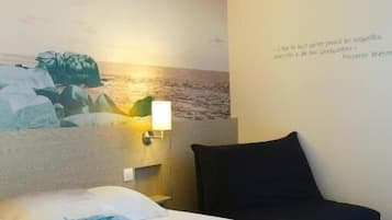 Una camera d'albergo con un letto, una lampada a muro e un quadro raffigurante una spiaggia.