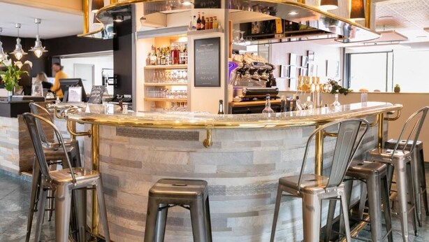 Zona de bar con una barra curva, taburetes y una variedad de botellas y vasos.