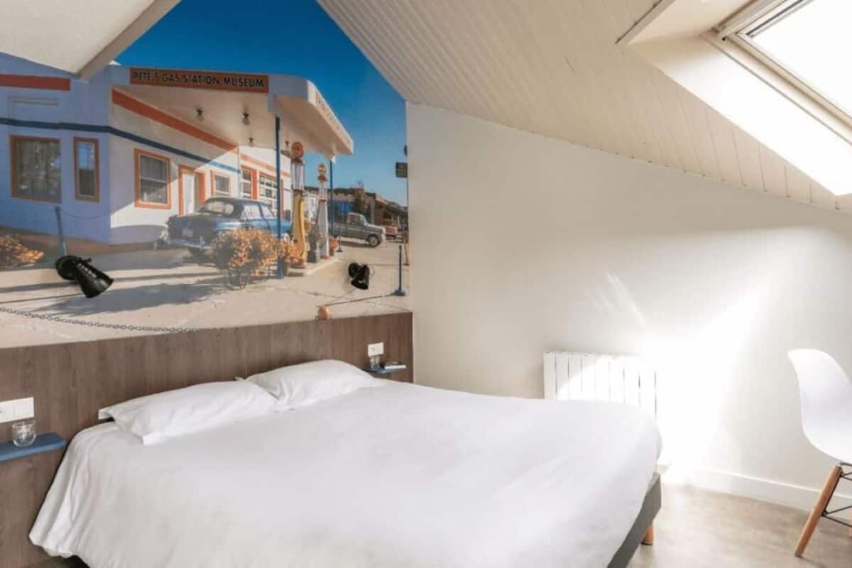 Hotelkamer met een bed, een stoel, een nachtkastje en een muurschildering van een benzinestation.