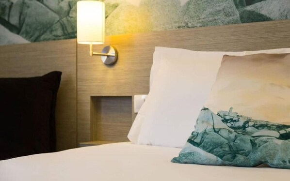 A neatly made bed with a white pillow and a green patterned pillow, a wall-mounted light, and a dark-colored pillow on the left. - Logis Hôtel Au Chêne Vert (Plerin)