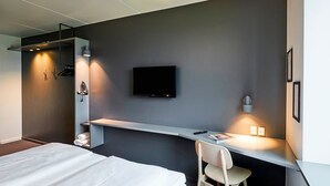Una habitación de hotel moderna con una cama, un escritorio con silla, un televisor de pantalla plana y un estante en la pared.
